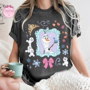 May include: Dark gray t-shirt featuring a framed image of Olaf from Frozen, with a blue bird, snowflakes, and a pink bow. Additional graphics include small snowmen and leaves. The shirt has a relaxed fit and short sleeves.