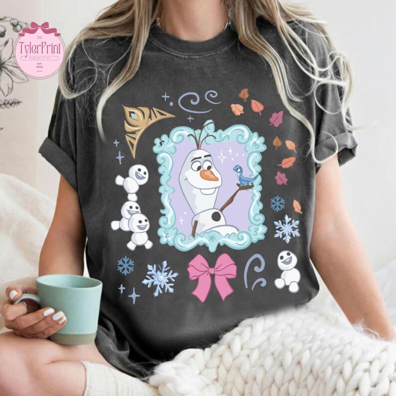 May include: Dark gray t-shirt featuring a framed image of Olaf from Frozen, with a blue bird, snowflakes, and a pink bow. Additional graphics include small snowmen and leaves. The shirt has a relaxed fit and short sleeves.