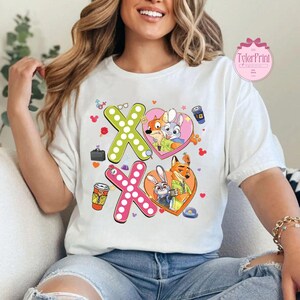 May include: White t-shirt featuring a colorful graphic design with the text "XO" and cartoon characters in heart-shaped frames. The design includes a fox and a bunny, along with coffee cups and other small icons. The shirt has the text "TylerPrint" on it.