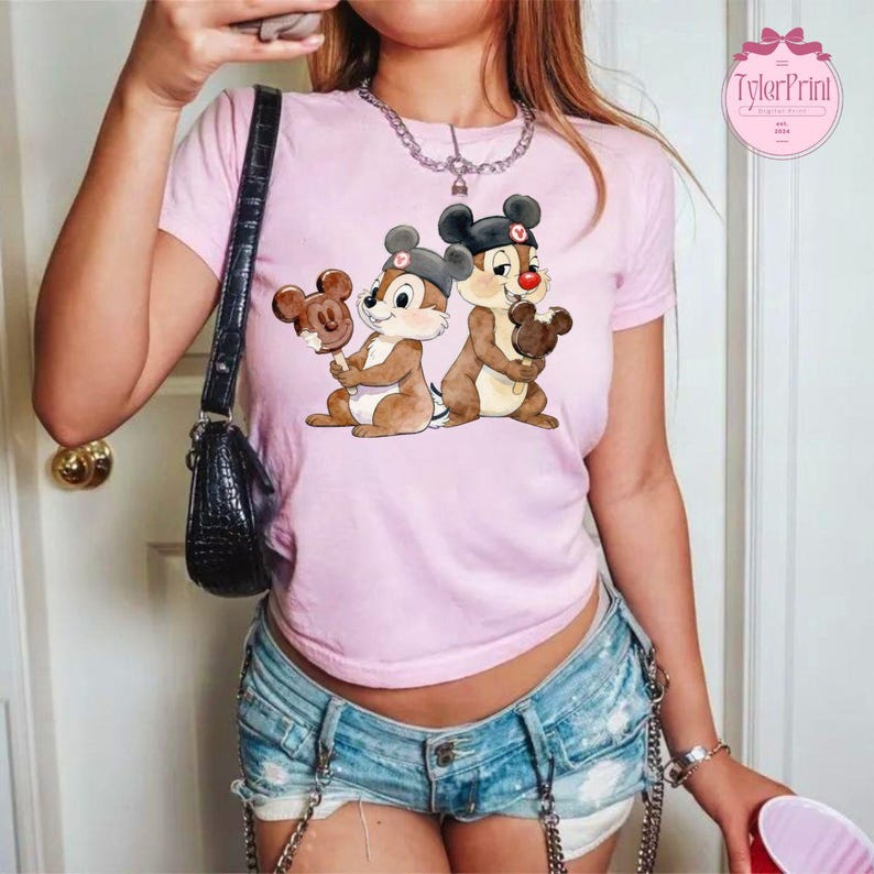 Puede incluir: Camiseta rosa claro con un gr&aacute;fico de dos ardillas listadas de dibujos animados sosteniendo golosinas con forma de Mickey Mouse. Las ardillas listadas son marrones y blancas, con sombreros negros. La camiseta tiene mangas cortas.