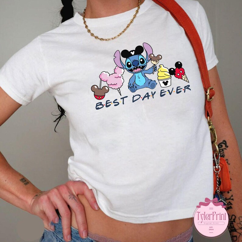 Pode incluir: Camiseta branca com um personagem Stitch azul segurando um cone de sorvete do Mickey Mouse. A camiseta tamb&eacute;m apresenta outros personagens da Disney e o texto "Best Day Ever".