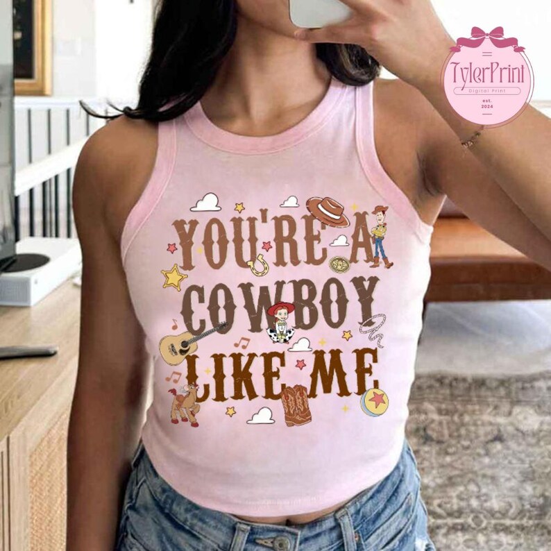 May include: A light pink sleeveless tank top featuring the brown text "YOU'RE A COWBOY LIKE ME" surrounded by colorful illustrations of a cowboy hat, a guitar, a horse, a boot, a lasso, stars, musical notes, and a cartoon character.