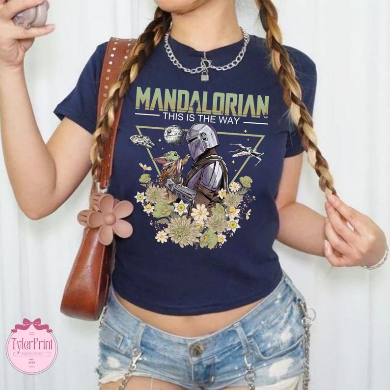 May include: Navy blue t-shirt featuring a graphic of The Mandalorian and Grogu surrounded by flowers, with the text "MANDALORIAN THIS IS THE WAY". The design includes a spaceship and the Death Star. A stylish tee for fans.