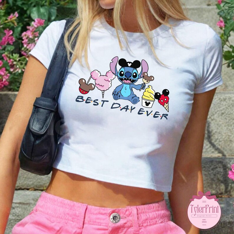 Pode incluir: Camiseta branca cropped com um personagem Stitch azul segurando um cone de sorvete do Mickey Mouse. A camiseta tem o texto "Best Day Ever" impresso.