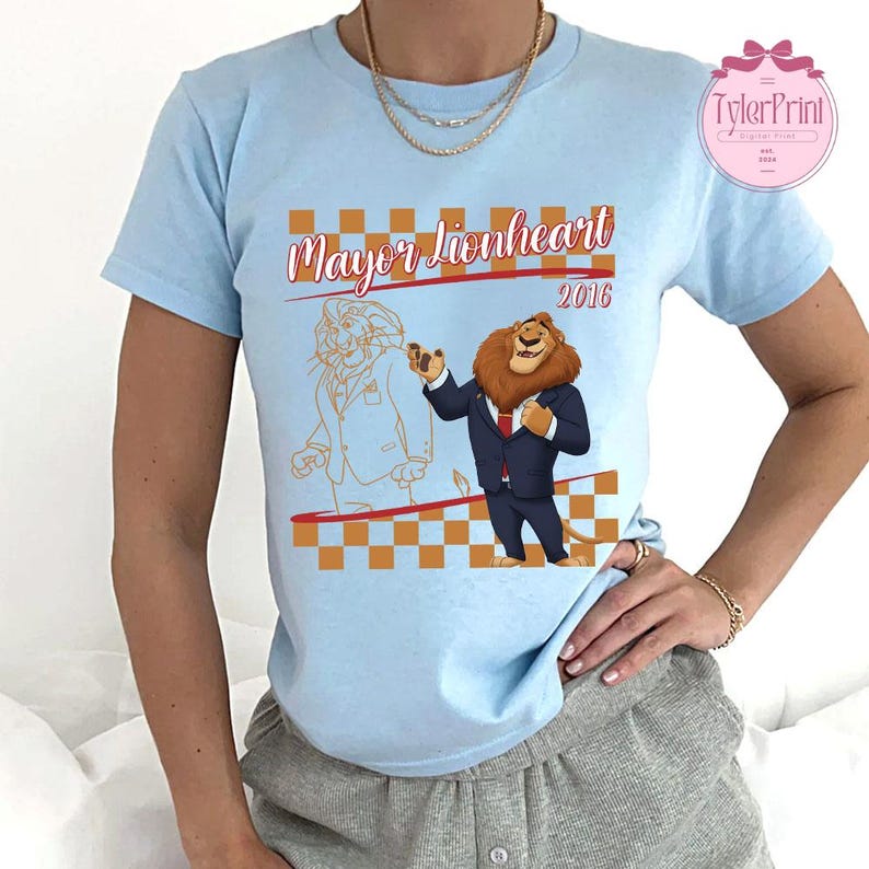 May include: Pale blue t-shirt with a cartoon lion design. The design includes the text "Mayor Lionheart 2016" and an orange and white checkered pattern. The lion is depicted in a suit. The shirt has short sleeves.
