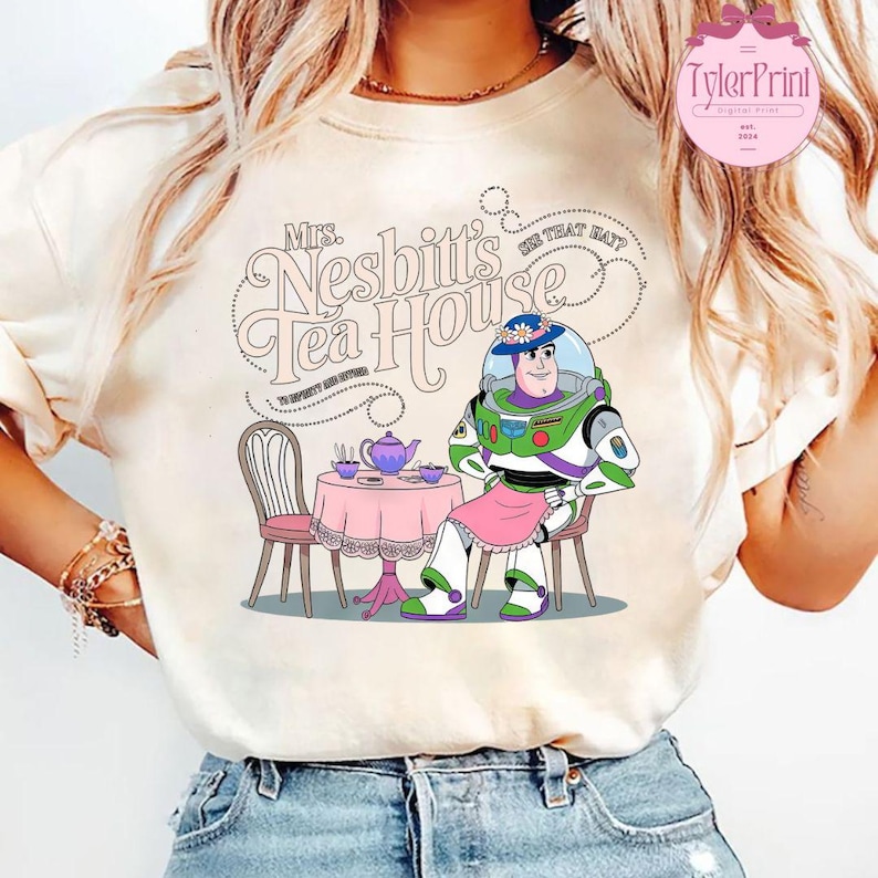 Może przedstawiać: Biały T-shirt z grafiką przedstawiającą Buzz Lightyeara z Toy Story przebranego za kobietę, siedzącego przy stole z czajnikiem i filiżankami do herbaty. Tekst na koszulce brzmi "Mrs. Nesbitt's Tea House".
