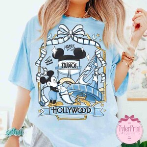 May include: Light blue t-shirt featuring a graphic of Mickey Mouse in front of a Hollywood Studios design. The design includes film strips, stars, and the words "Mickey Studios" and "Hollywood". The shirt has short sleeves.
