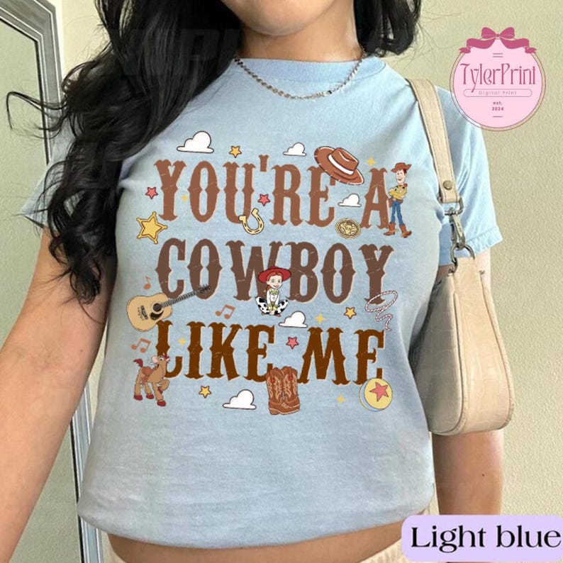 May include: A light blue t-shirt featuring brown text that reads YOU'RE A COWBOY LIKE ME. The graphic includes illustrations of a cowboy hat, a guitar, a lasso, a horse, cowboy boots, a ball, and two small cartoon characters in western attire.