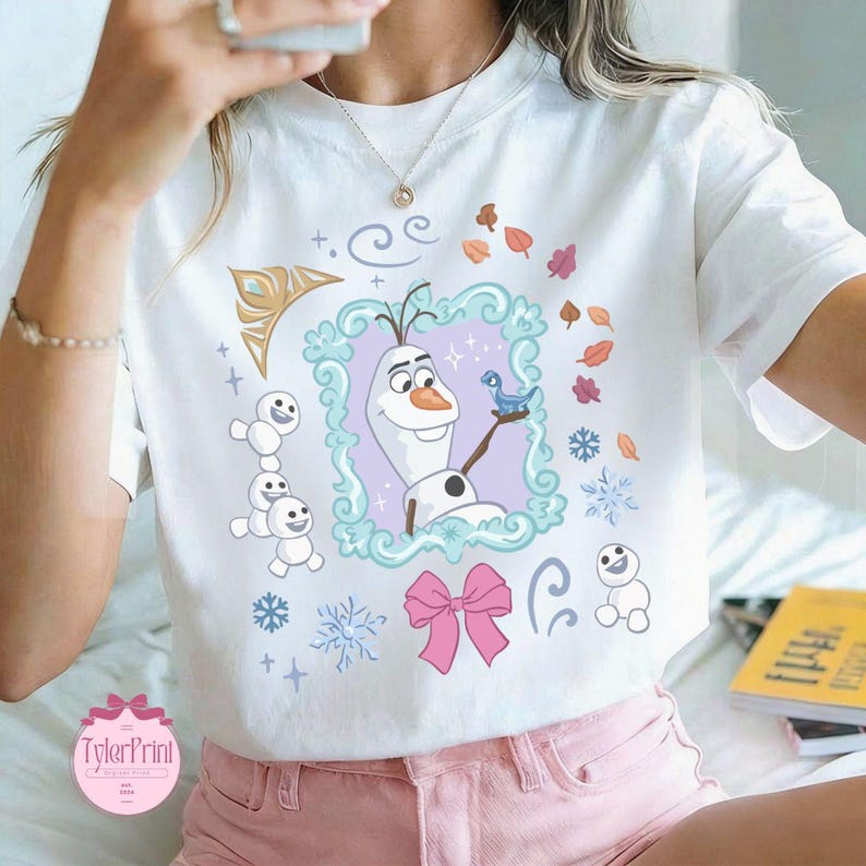 May include: White t-shirt featuring a colorful graphic of Olaf from Frozen, surrounded by snowflakes, leaves, and a pink bow. The design includes the text "TylerPrint" in a pink circle. The shirt is being worn.
