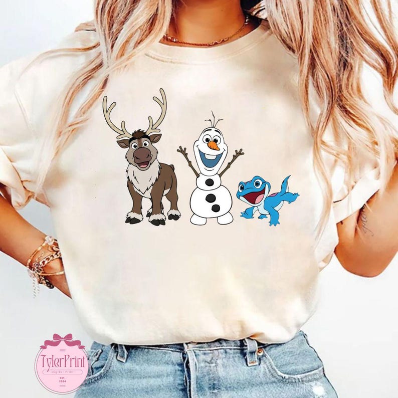 Peut inclure: T-shirt cr&egrave;me avec des personnages de dessins anim&eacute;s du film La Reine des Neiges : un renne, un bonhomme de neige et un l&eacute;zard bleu. Le renne est marron avec de grands bois. Le bonhomme de neige est blanc avec un visage souriant.