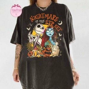 K&ouml;nnte beinhalten: Schwarzes T-Shirt mit einer farbenfrohen Grafik aus "Nightmare Before Christmas". Das Design zeigt Jack Skellington, Sally und den Text "Nightmare on Main Street". Das Shirt hat einen Vintage-Look mit leicht verblichenen Farben.