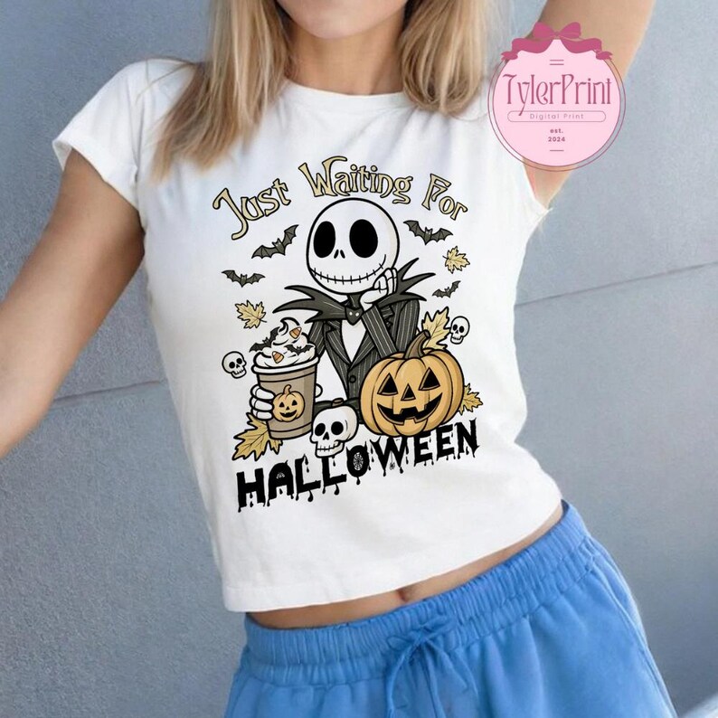 Puede incluir: Camiseta blanca con un dise&ntilde;o tem&aacute;tico de Halloween. El dise&ntilde;o incluye un personaje de esqueleto de dibujos animados, una calabaza tallada, caf&eacute;, murci&eacute;lagos y el texto "Just Waiting For Halloween". El dise&ntilde;o es negro, naranja y marr&oacute;n.