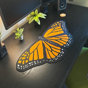 May include: A desk mat shaped like a monarch butterfly wing with vibrant orange and black patterns, featuring white spots along the edges and the word "PROTO" printed in a dark gray font on the central section of the butterfly wing design.