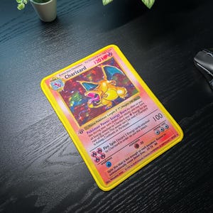 以下が含まれることがあります： 黄色い縁取りの、Charizardのポケモンカード。カードには、「Charizard」と「120 HP」の文字が入った、ドラゴンに似た生き物のカラフルなイラストが描かれています。カードは濃い木材の表面にあります。