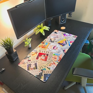 Może przedstawiać: Czarny stół z dwoma monitorami komputerowymi, klawiaturą i podkładką pod mysz. Podkładka pod mysz przedstawia kolorowy kolaż plakatów "Wanted" z różnymi postaciami z anime. Widoczne są również małe rośliny doniczkowe i zielone krzesło.