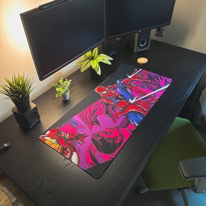 May include: A black desk with two computer monitors, speakers, and a mousepad featuring a comic book illustration of Spider-Man and Iron Man. The desk also has small potted plants and a green chair.