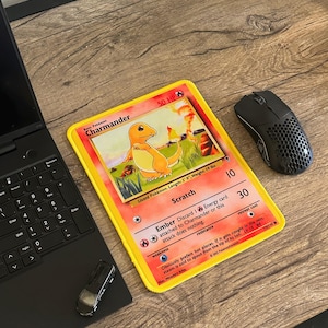 Può includere: Una carta collezionabile Pokémon Charmander gialla e arancione con il testo "Charmander" e "50 HP". La carta è su una scrivania di legno accanto a un laptop nero, un mouse e un piccolo dispositivo nero.