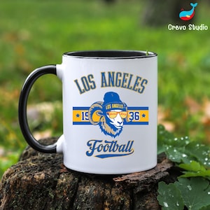 Puede incluir: Taza de cerámica blanca con asa y borde negros. La taza presenta el texto "LOS ANGELES" en azul, junto con un gráfico de carnero con gorro azul y gafas de sol. El texto "Football" también se muestra en azul.