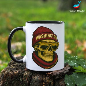 May include: White ceramic mug with a black handle and rim. The mug features a skull design wearing a red beanie with "WASHINGTON" printed on it, sunglasses, and a red scarf. The words "Washington Football" are printed on the mug.