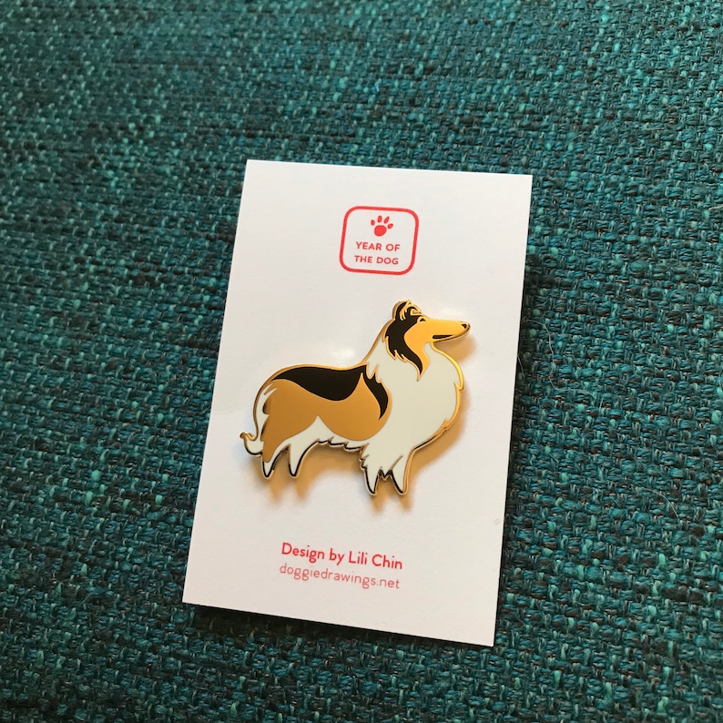 Puede incluir: Pin de esmalte dorado con un perro Collie de pelo negro, blanco y dorado. El pin dice "A&ntilde;o del Perro" y "Dise&ntilde;o de Lili Chin, doggiedrawings.net".