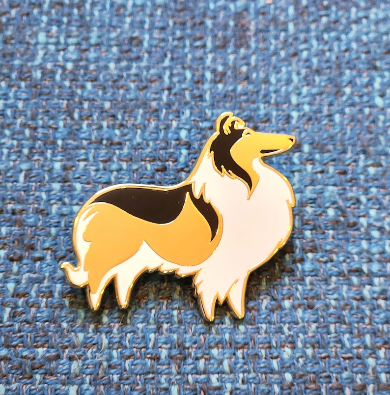 Puede incluir: Un pin de esmalte dorado con un perro Collie de dibujos animados con pelo negro, blanco y marr&oacute;n. El perro est&aacute; de pie sobre sus cuatro patas y mirando hacia la derecha.