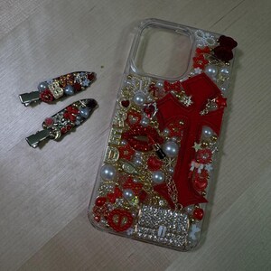 May include: A clear phone case decorated with red and gold embellishments, including a large red cross, pearls, and rhinestones. The case has a cutout for the camera lens.