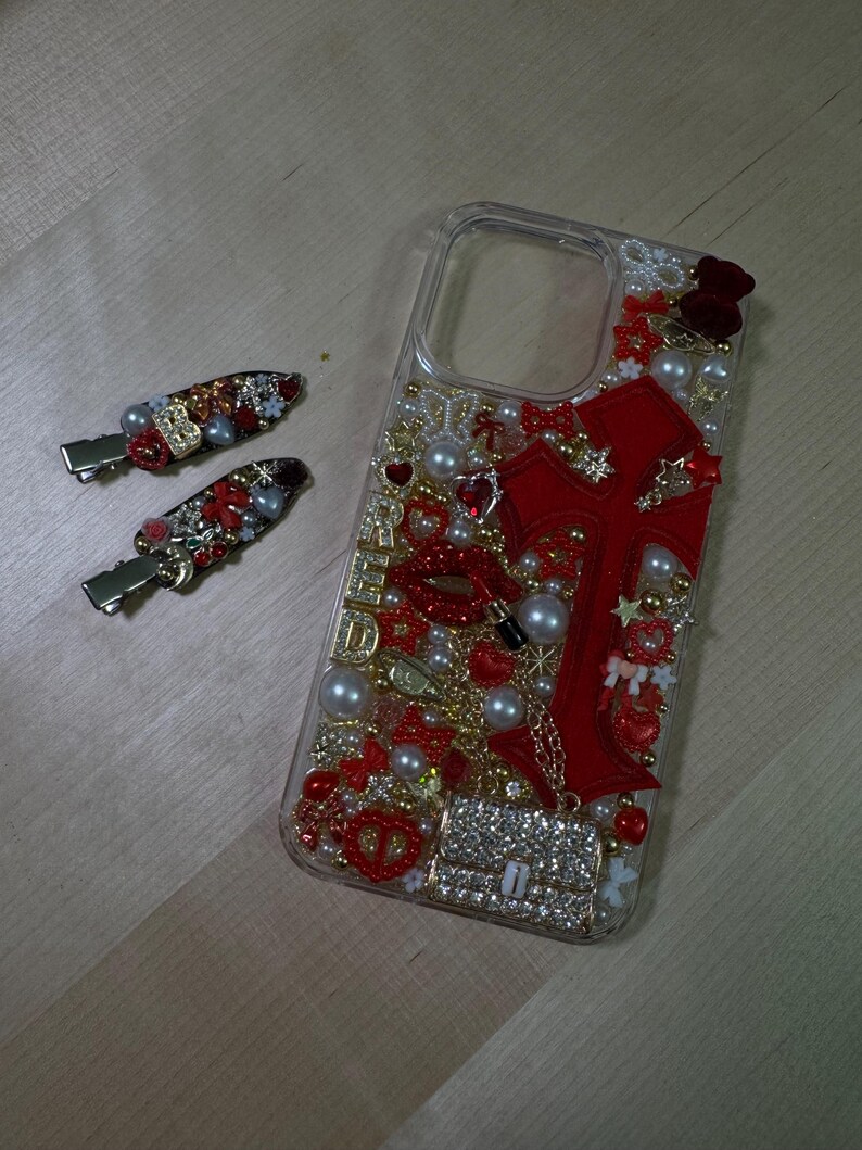 May include: A clear phone case decorated with red and gold embellishments, including a large red cross, pearls, and rhinestones. The case has a cutout for the camera lens.