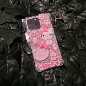 May include: A pink Hello Kitty phone case with a clear background and glitter. The case features a 3D Hello Kitty figure, a checkered design, and other embellishments.