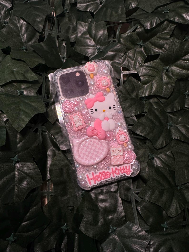 May include: A pink Hello Kitty phone case with a clear background and glitter. The case features a 3D Hello Kitty figure, a checkered design, and other embellishments.