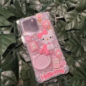 May include: A clear phone case with a pink Hello Kitty design. The case is decorated with rhinestones, glitter, and 3D Hello Kitty characters. The case has a pink and white checkered pattern on the back.