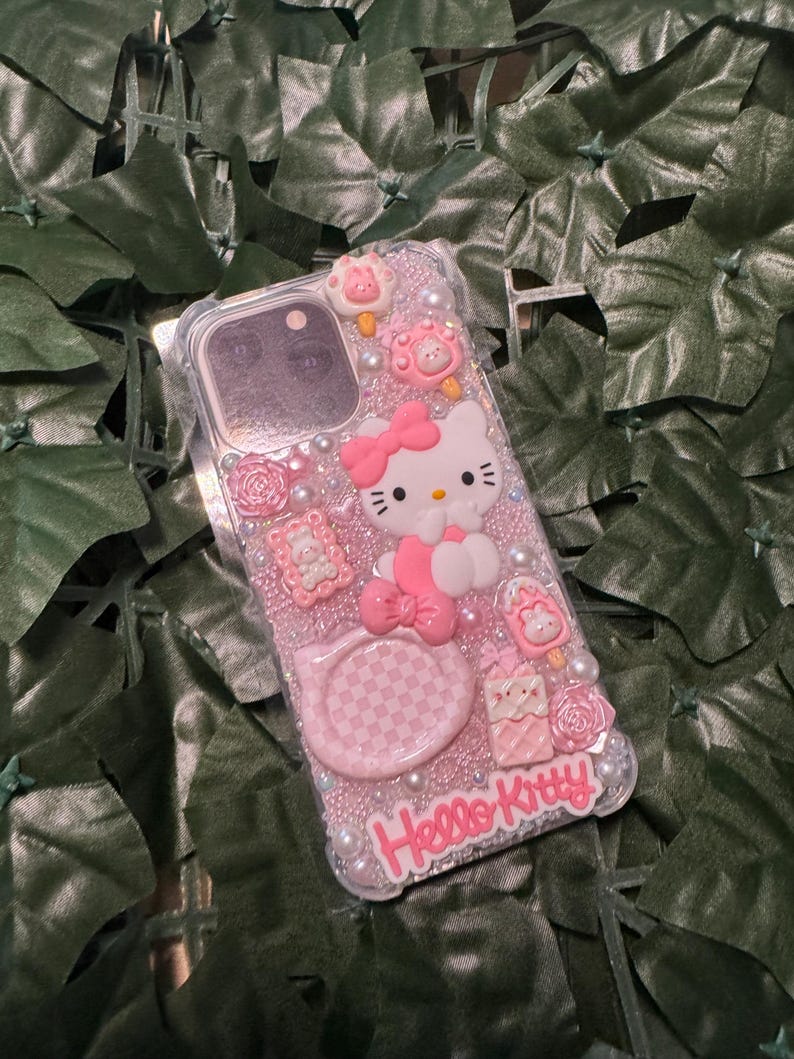 May include: A clear phone case with a pink Hello Kitty design. The case is decorated with rhinestones, glitter, and 3D Hello Kitty characters. The case has a pink and white checkered pattern on the back.