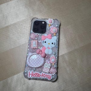 May include: A clear phone case with a pink and white Hello Kitty design. The case has glitter and 3D embellishments, including a Hello Kitty character, a bow, and flowers.