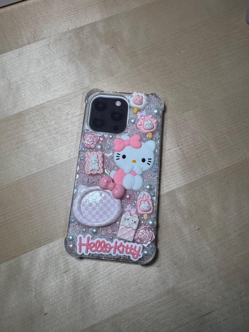 May include: A clear phone case with a pink and white Hello Kitty design. The case has glitter and 3D embellishments, including a Hello Kitty character, a bow, and flowers.