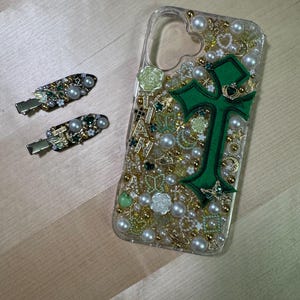 May include: A clear phone case decorated with green, gold, and white embellishments. The case features a large green cross with gold accents. The case is filled with small pearls, rhinestones, and other decorative elements.
