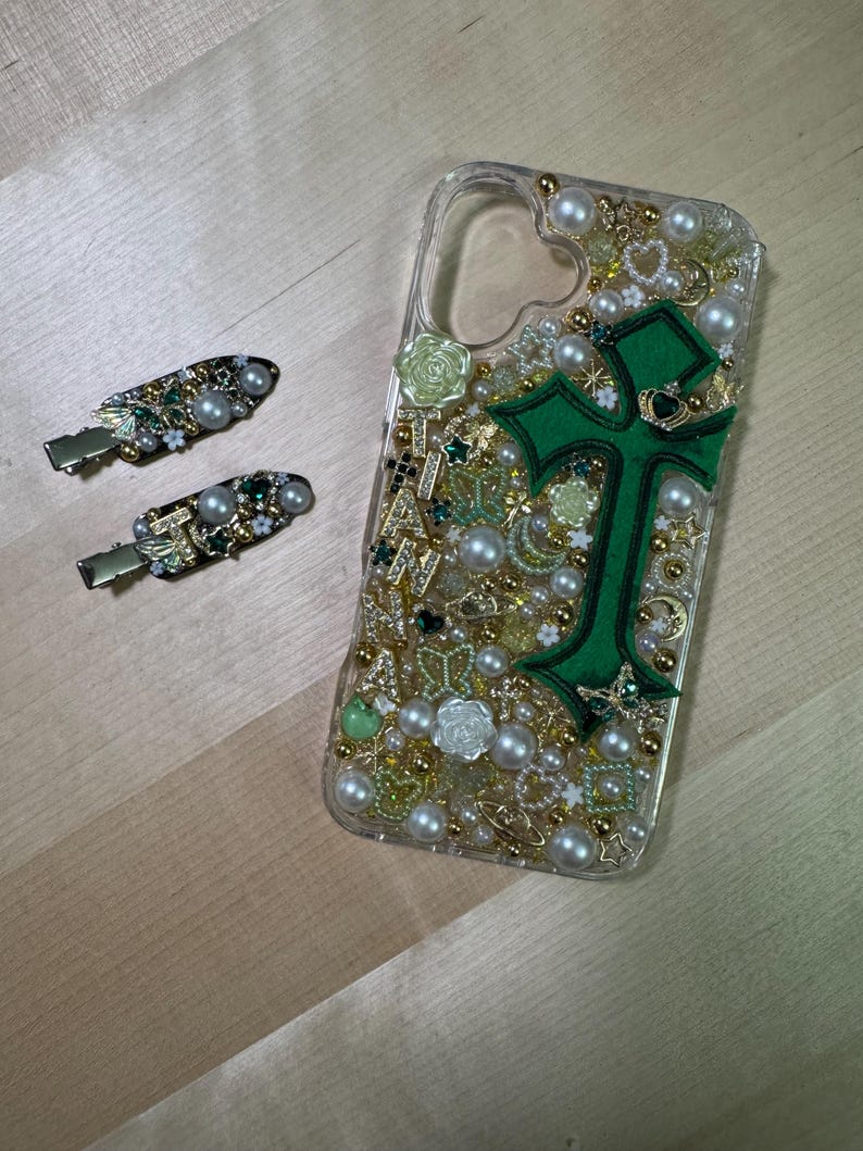 May include: A clear phone case decorated with green, gold, and white embellishments. The case features a large green cross with gold accents. The case is filled with small pearls, rhinestones, and other decorative elements.