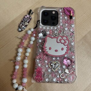 May include: A clear phone case decorated with pink and white pearls, rhinestones, and charms. The case features a Hello Kitty charm, a pink heart charm, and a safety pin charm. A pink and white beaded phone strap is attached to the case.