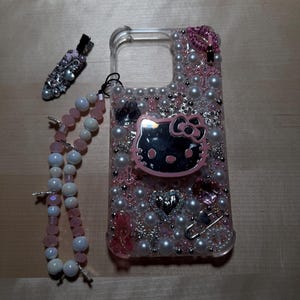 May include: A clear phone case decorated with pink and white pearls, rhinestones, and a Hello Kitty charm. The case also has a pink and white beaded strap.