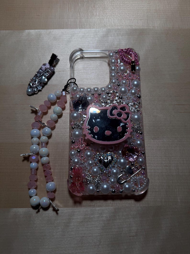 May include: A clear phone case decorated with pink and white pearls, rhinestones, and a Hello Kitty charm. The case also has a pink and white beaded strap.