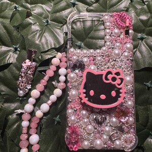 May include: A pink and white phone case with a Hello Kitty design, adorned with pearls, rhinestones, and other embellishments. The case also includes a pink and white beaded phone strap.