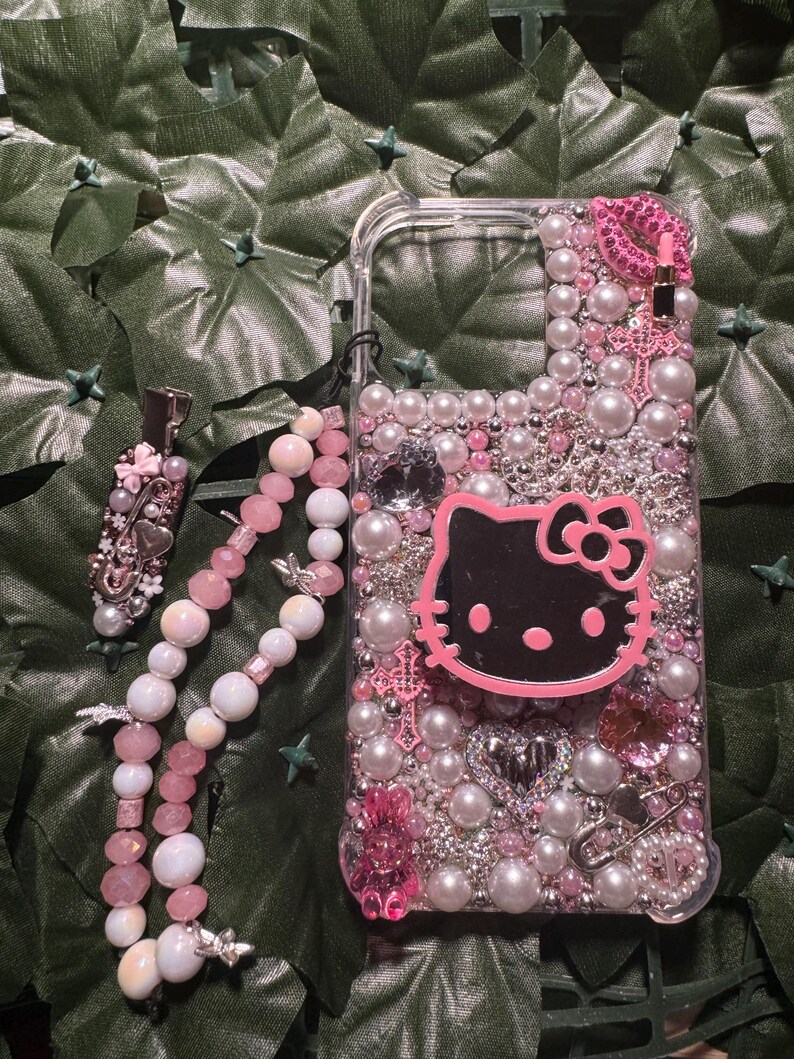 May include: A pink and white phone case with a Hello Kitty design, adorned with pearls, rhinestones, and other embellishments. The case also includes a pink and white beaded phone strap.
