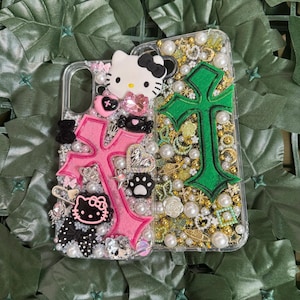 Pode incluir: Duas capas transparentes para telem&oacute;vel decoradas com strass, glitter e pingentes. Uma capa apresenta uma cruz rosa e uma personagem Hello Kitty, enquanto a outra apresenta uma cruz verde.