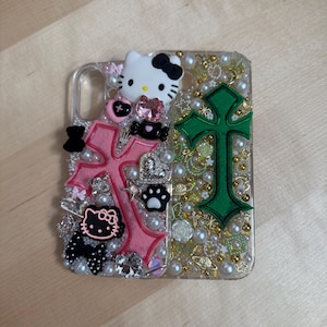 Pode incluir: Uma capa transparente para telefone decorada com cruzes rosa e verde, personagens Hello Kitty, p&eacute;rolas e outros enfeites.