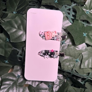 May include: Two black hair clips decorated with pink, white, and silver embellishments. One clip features a pink and white cake with a small pink bunny on top. The other clip features a black star and a pink bow.