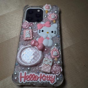 May include: A clear phone case with a pink Hello Kitty design. The case is decorated with glitter, pearls, and 3D embellishments, including a Hello Kitty figurine, a pink bow, and a checkered pattern.