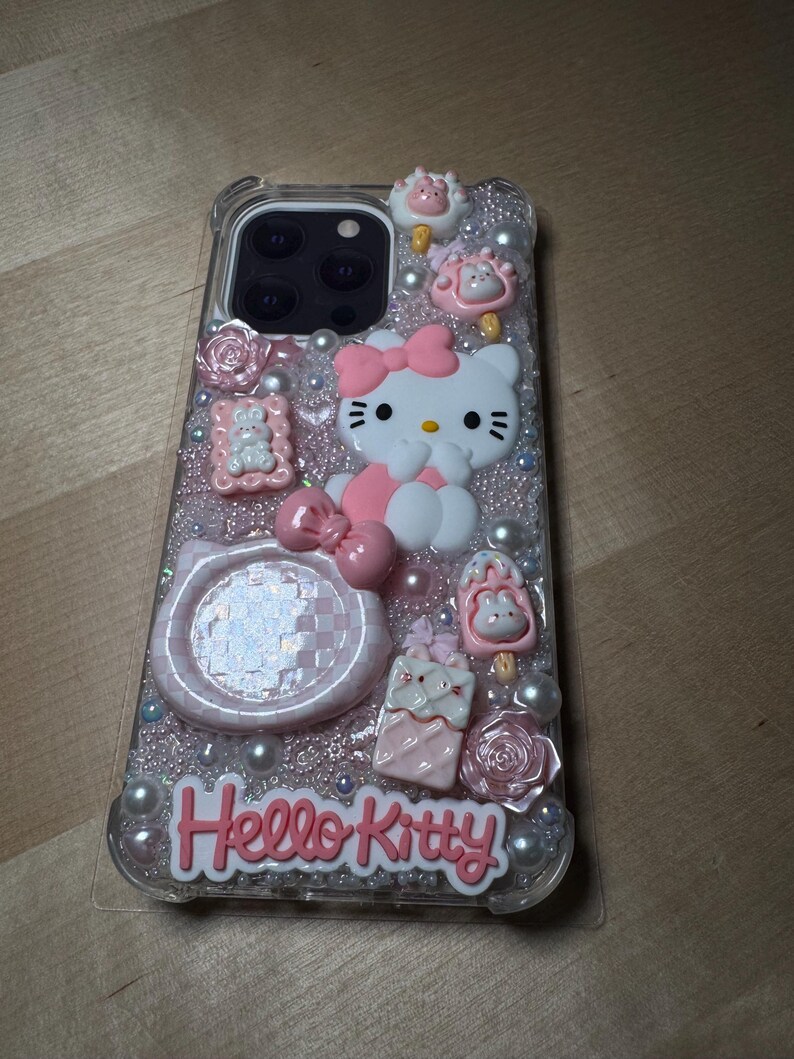May include: A clear phone case with a pink Hello Kitty design. The case is decorated with glitter, pearls, and 3D embellishments, including a Hello Kitty figurine, a pink bow, and a checkered pattern.