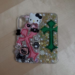 Pode incluir: Uma capa transparente para telefone com um design de cruz rosa e verde. A capa &eacute; decorada com strass, p&eacute;rolas e pingentes, incluindo um personagem Hello Kitty.