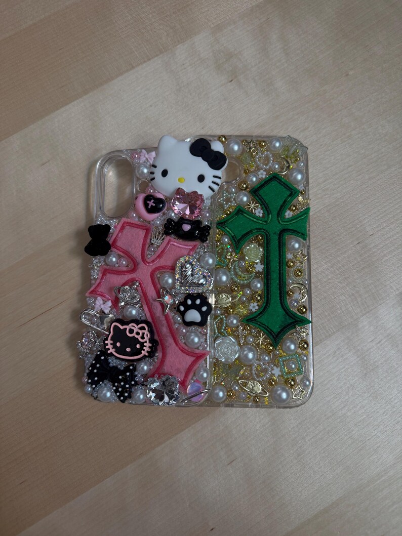 Pode incluir: Uma capa transparente para telefone com um design de cruz rosa e verde. A capa &eacute; decorada com strass, p&eacute;rolas e pingentes, incluindo um personagem Hello Kitty.