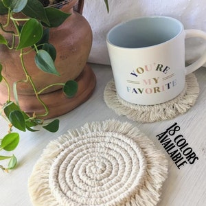 May include: A macrame coaster with a spiral design and fringed edges sits on a white surface. A mug with the text "YOU'RE MY FAVORITE" rests on a similar coaster. A terracotta pot with a green plant is in the background. The text "18 COLORS AVAILABLE" is also visible.