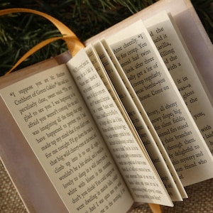May include: A miniature book with cream-colored pages featuring printed black text from Anne of Green Gables. The book is open, displaying multiple pages fanned out, and includes a thin orange ribbon attached to the spine for use as a bookmark.