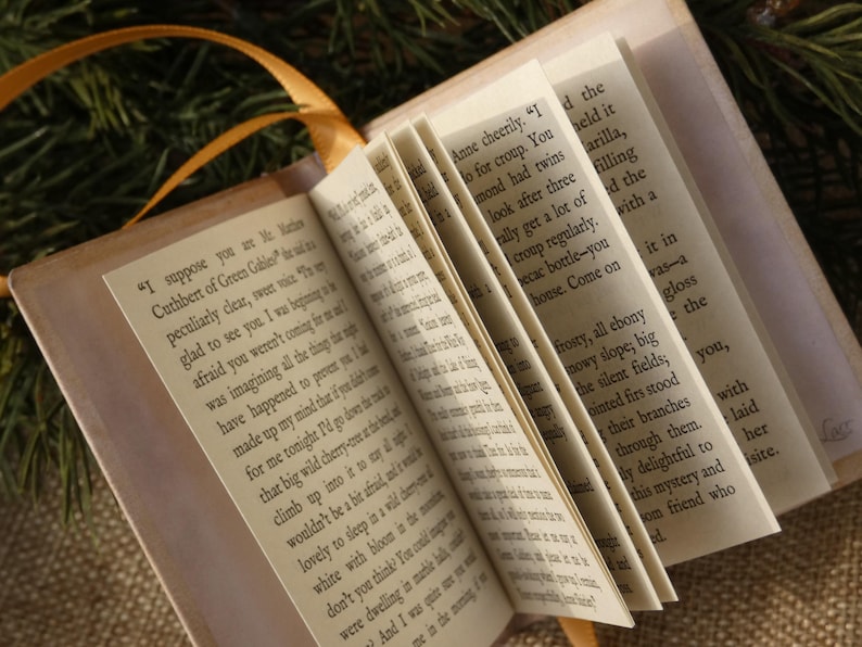 May include: A miniature book with cream-colored pages featuring printed black text from Anne of Green Gables. The book is open, displaying multiple pages fanned out, and includes a thin orange ribbon attached to the spine for use as a bookmark.